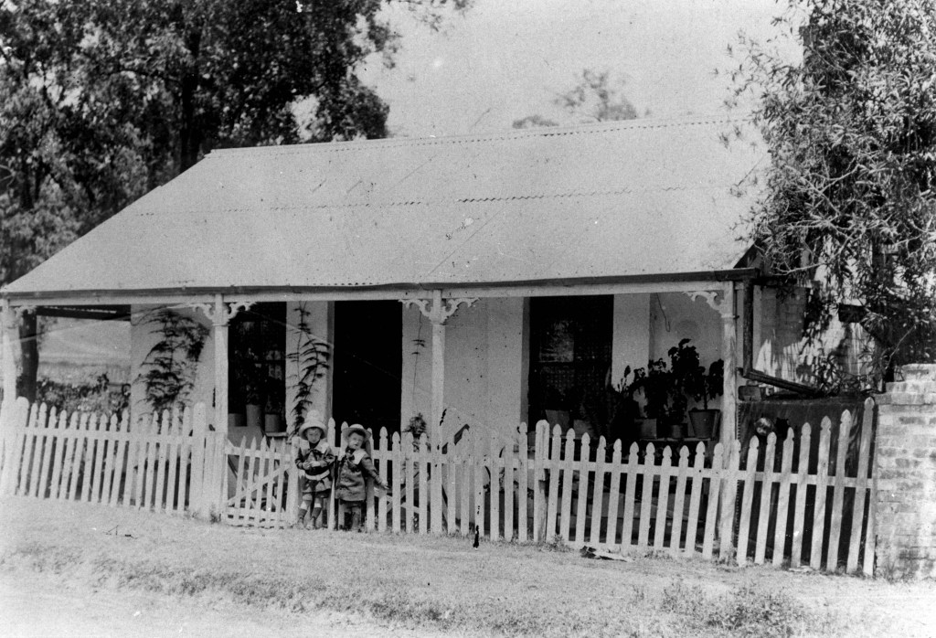 Two tiny sailor suited young lads standing at the entrance of a charming whitewashed cottage in Alderley about 1910. Cottage enclosed with a paling fence stood on South Pine Road at Alderley.